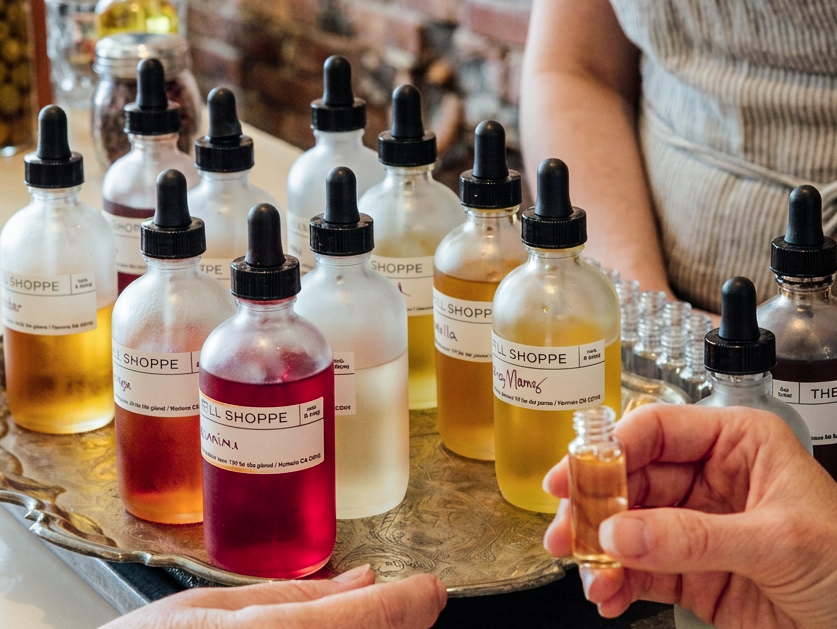 Hand holding a small Refill Shoppe essential oil dropper bottle next to a tray of labeled oil blends in various colors