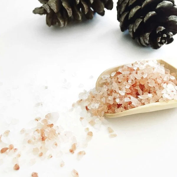 Wooden scoop of pink Himalayan bath salts with pine cones on a white background