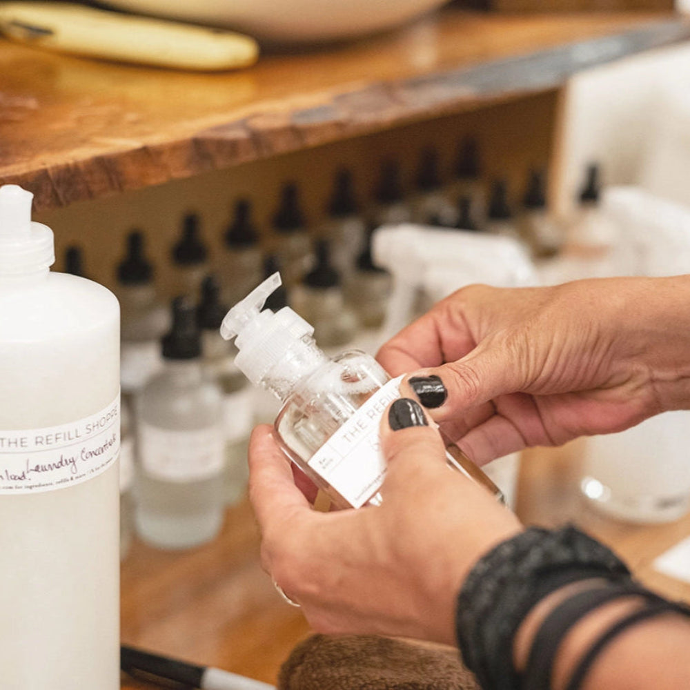 Staff putting label on a bottle in The Refill Shoppe with various bottles on a wooden shelf.