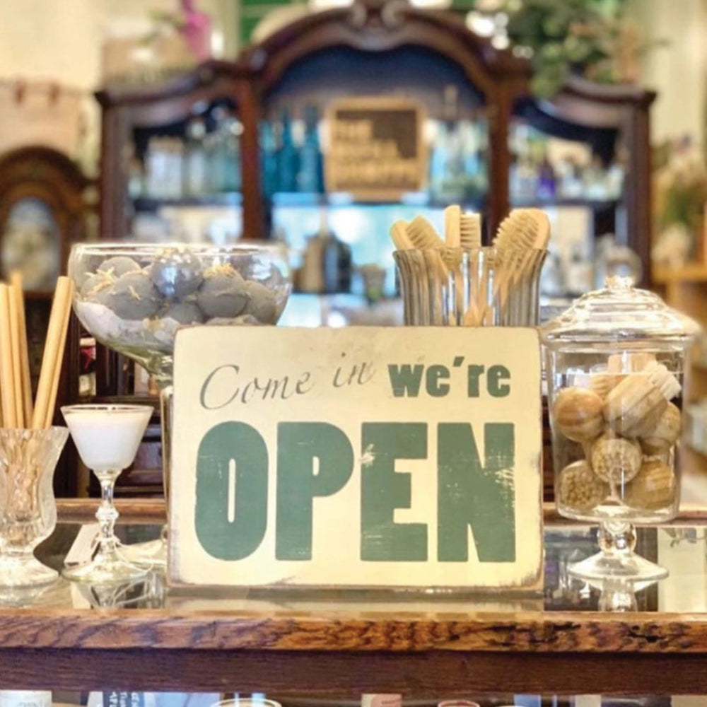A sign that says "Come in, we're open" sitting on a wooden counter with various products in The Refill Shoppe
