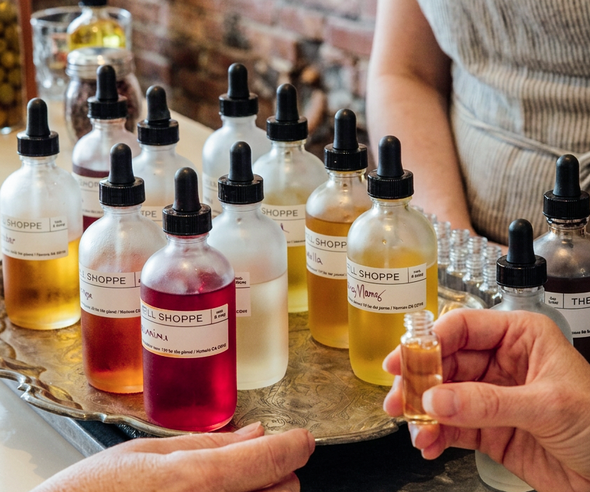 Hand holding a small Refill Shoppe essential oil dropper bottle next to a tray of labeled oil blends in various colors