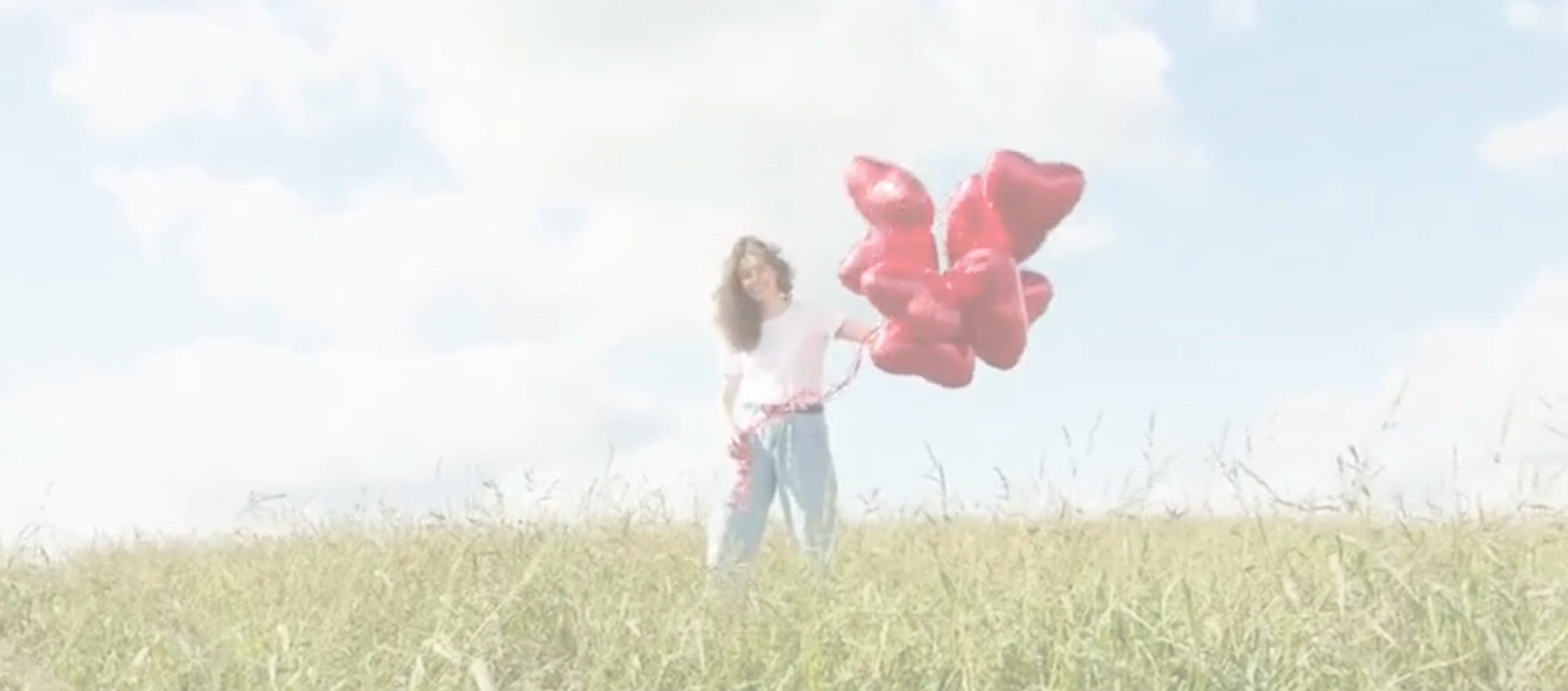 Woman standing in a green field with a bunch of red heart balloons against a blue sky
