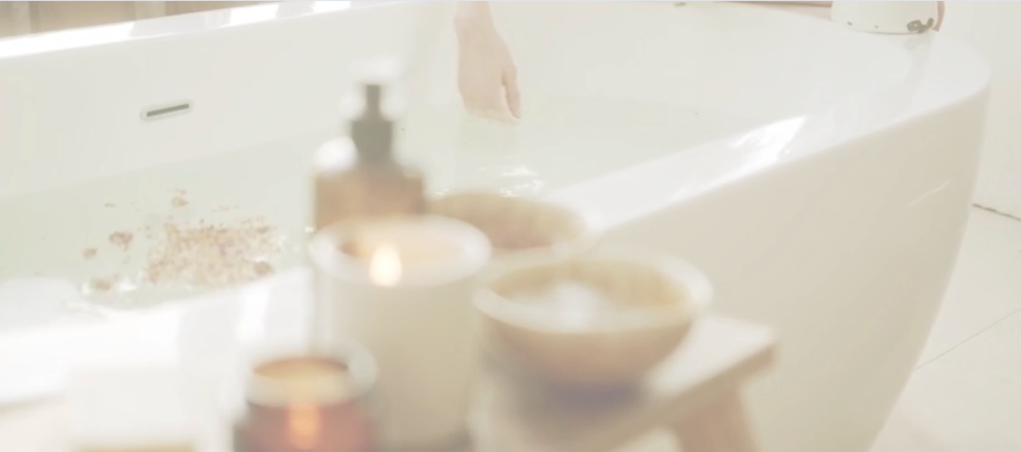 Blurred bath products in the foreground and a bathtub in the background with flower petals in the water