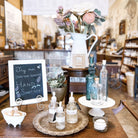 Hand sanitizer bottles on a table with a sign and decorative items and glass bottles in a store setting