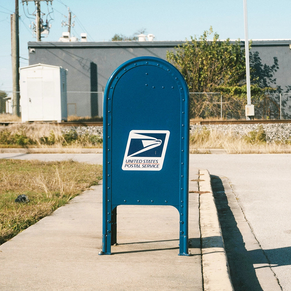 Blue United States Postal Service mailbox on a sidewalk with buildings and a clear sky in the background