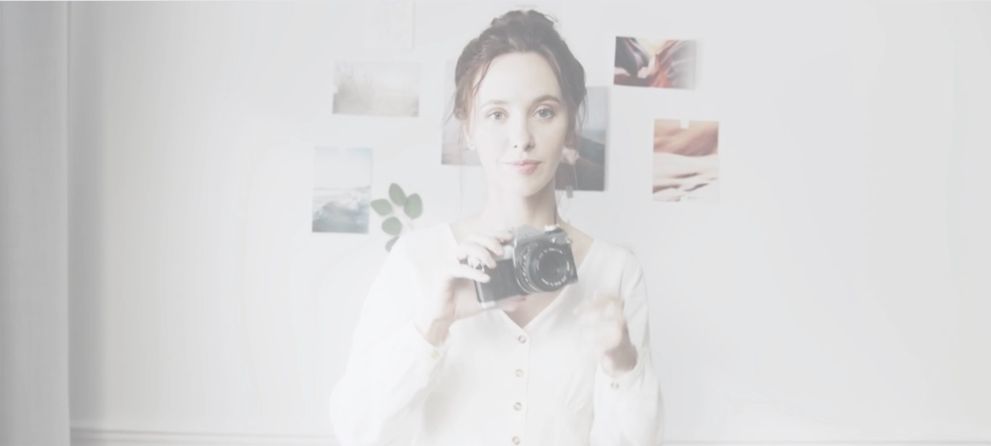 Woman wearing a white dress holding a camera with photographs on the white wall behind her.